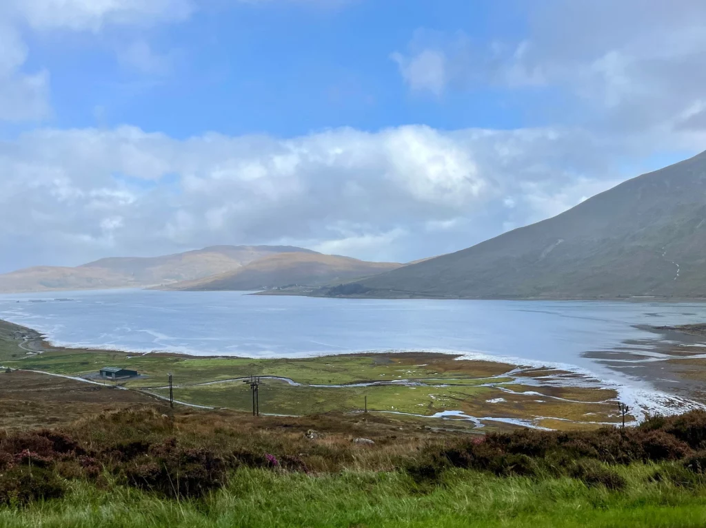 Eine weite Meeresbucht oder ein großer See, umgeben von hohen, kahlen Bergen unter einem leicht bewölkten blauen Himmel. Im Vordergrund erstreckt sich eine grüne Küstenebene mit kleinen Wasserläufen, die in das ruhige, silbrig glänzende Wasser münden.
