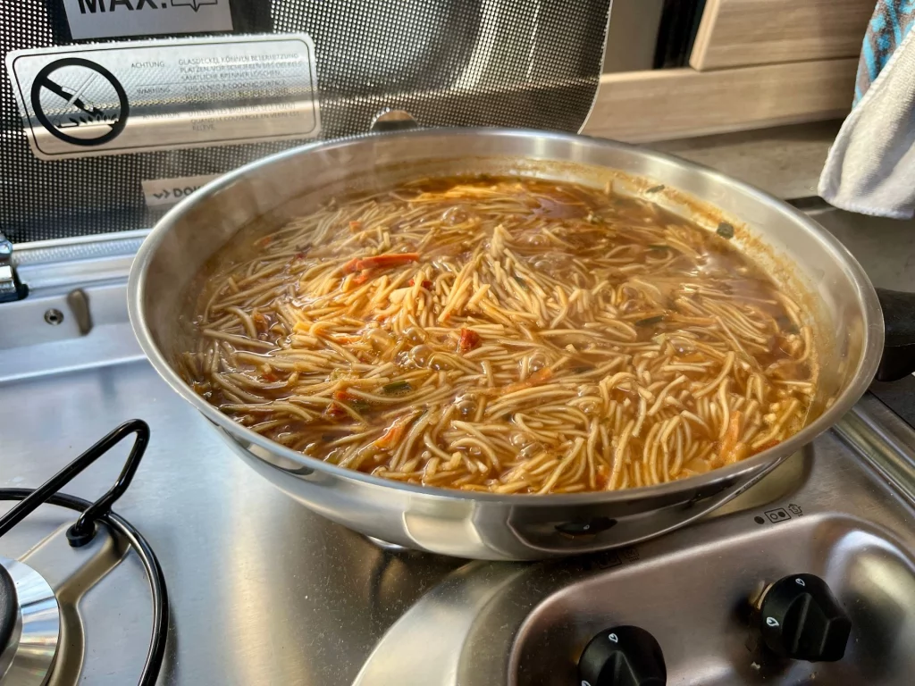 Eine Nahaufnahme einer dampfenden Pfanne mit einer herzhaften Nudelsuppe oder einem Eintopf, der gerade auf einem Gasherd im Camper zubereitet wird.
