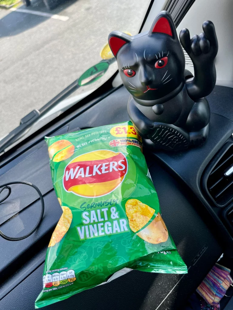 Auf dem Armaturenbrett eines Autos liegt eine grüne Tüte „Walkers Salt & Vinegar“ Chips neben einer schwarzen Winkekatze (Maneki-neko), die das „Rock on“-Handzeichen macht.