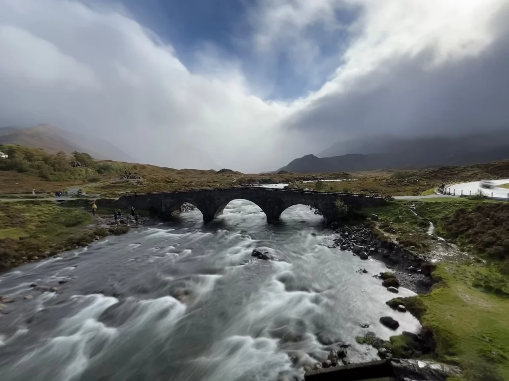 Dramatische Panoramaaufnahme der dreibogigen Sligachan Old Bridge über einem schnell fließenden Fluss, im Hintergrund die nebelverhangenen Cuillin-Berge.