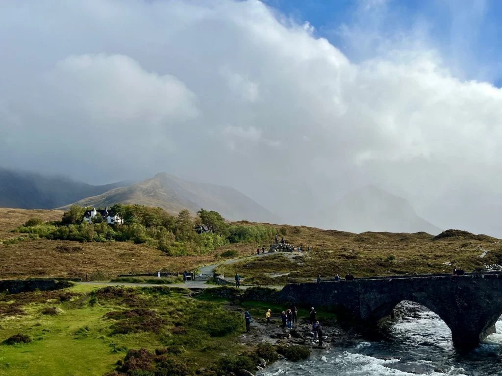 Blick über eine hügelige Graslandschaft unter dramatisch bewölktem Himmel mit der Silhouette der Sligachan Bridge im Hintergrund.
