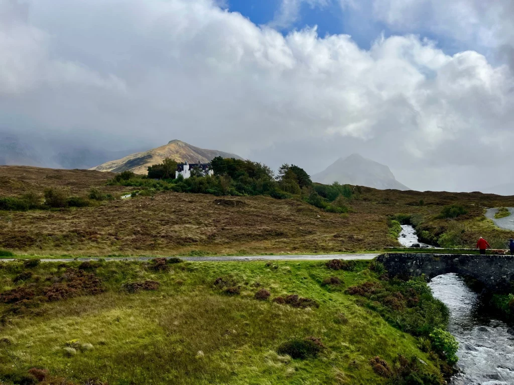 Eine sanfte Hügellandschaft mit herbstlichem Gras und einem kleinen weißen Haus in der Ferne unter grauem Himmel.