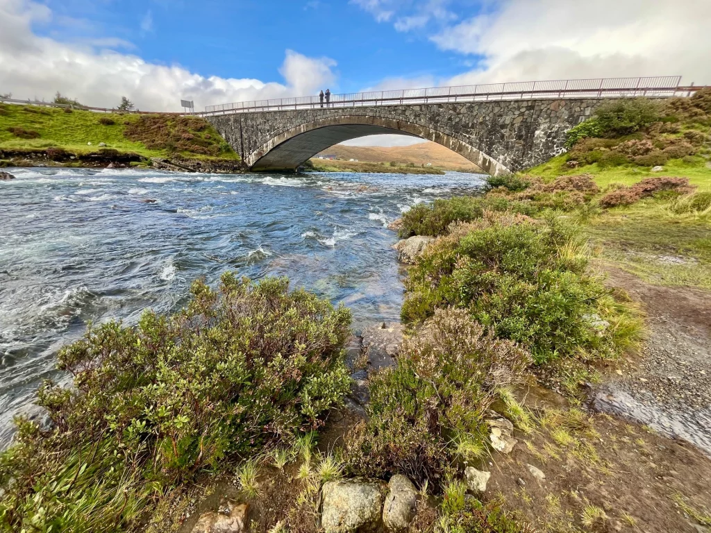 Seitliche Ansicht der alten Steinbrücke, die sich über den rauschenden Sligachan River spannt, umgeben von herbstlicher Vegetation.