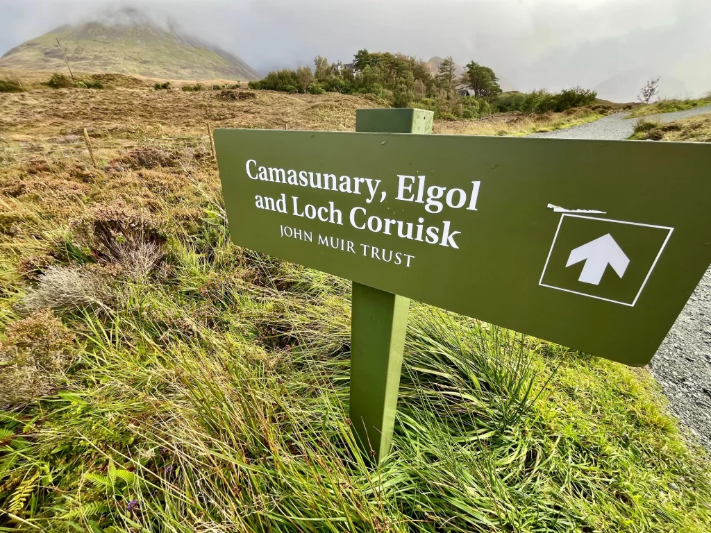 Ein grünes Hinweisschild aus Holz mit der Aufschrift „Cuillinary, Elgol and Loch Coruisk“, das in einer feuchten Graslandschaft steht.