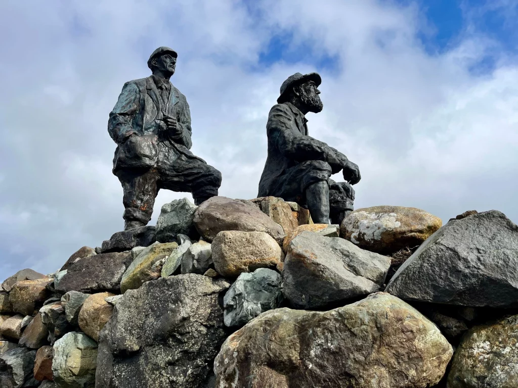 Bronzestatue von zwei Männern in historischer Bergsteigerkleidung (Norman Collie und John MacKenzie), die auf einem Sockel aus großen Natursteinen sitzen und in die Ferne blicken.