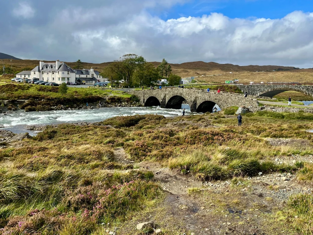 Blick über den Sligachan River auf das Sligachan Hotel und die alte Steinbrücke, eingebettet in die raue Hügellandschaft von Skye.