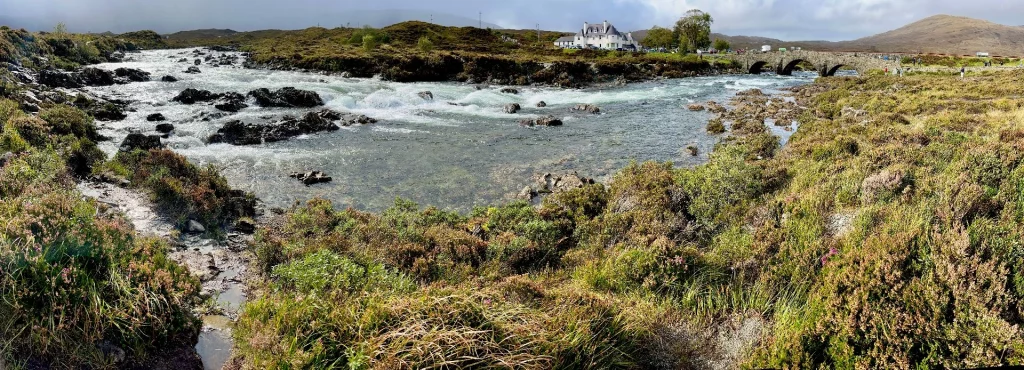 Weite Panoramaaufnahme des wilden Sligachan Rivers mit Stromschnellen; im Hintergrund sind das weiße Hotelgebäude, die Steinbrücke und die Ausläufer der Cuillin-Berge zu sehen.