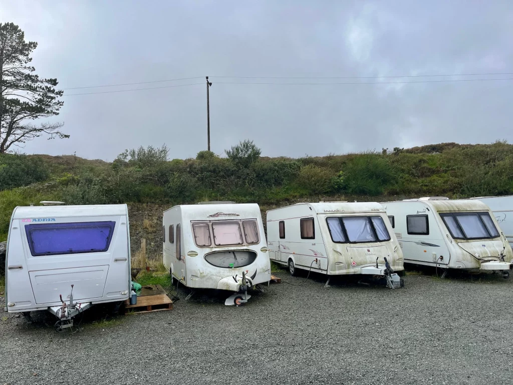 Vier ältere, weiße Wohnwagen stehen nebeneinander auf einem grauen Schotterplatz vor einer grünen Böschung unter einem trüben, grauen Himmel.