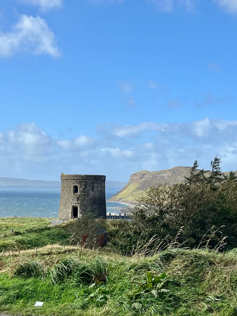 Ein historischer Steinturm (Uig Tower) auf einem grünen Hügel über dem Meer unter blauem Himmel.