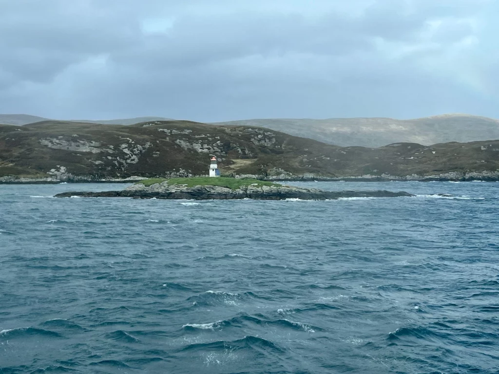 Ein kleiner weißer Leuchtturm auf einer felsigen Insel im Meer, dahinter die kargen, grünen Hügel der Isle of Harris.