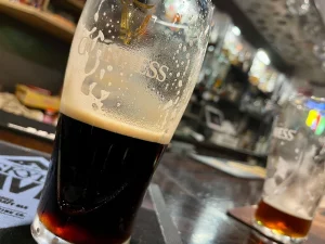 Nahaufnahme eines halb ausgetrunkenen Glases Guinness mit cremigem Schaum auf einem dunklen Tresen in einem Pub in Uig; im unscharfen Hintergrund sind die Bar-Armaturen und ein weiteres Glas zu erkennen.