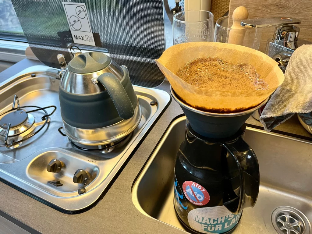 Innenansicht einer Kastenwagen-Küche; auf dem Gasherd steht ein faltbarer Wasserkocher, daneben ein Kaffeefilter auf einer Thermoskanne in der Spüle.