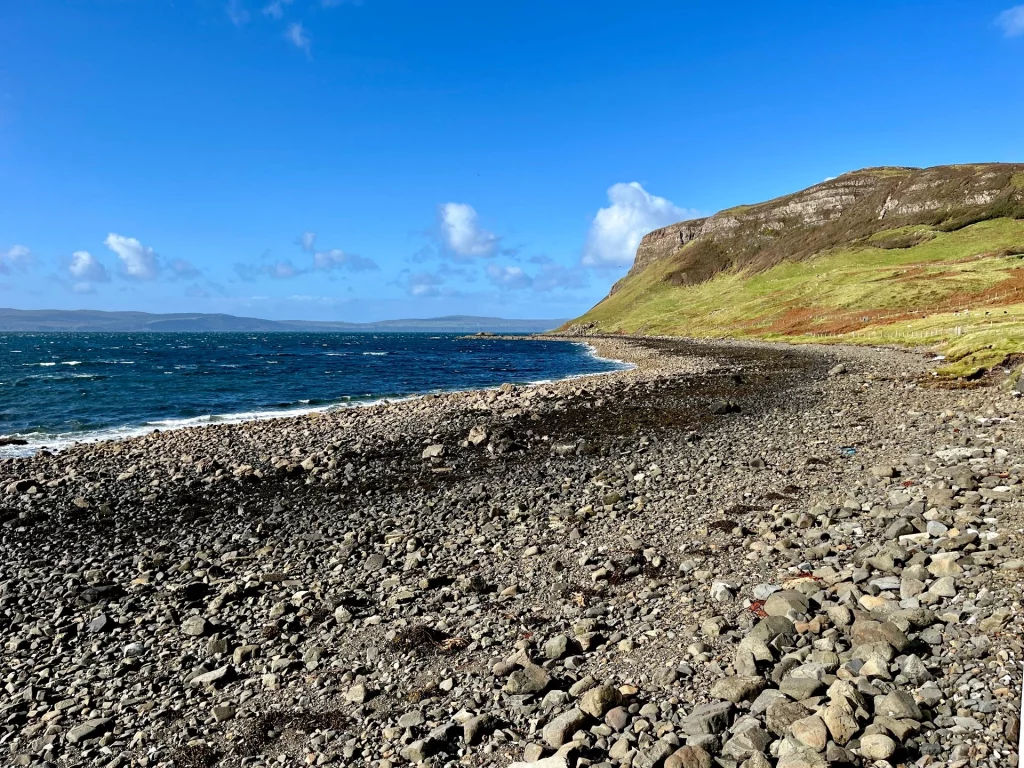 Ein weiter Blick über einen grauen Steinstrand an der Küste von Skye; im Hintergrund eine steile grüne Klippe und das tiefblaue Meer unter einem weiten Himmel.