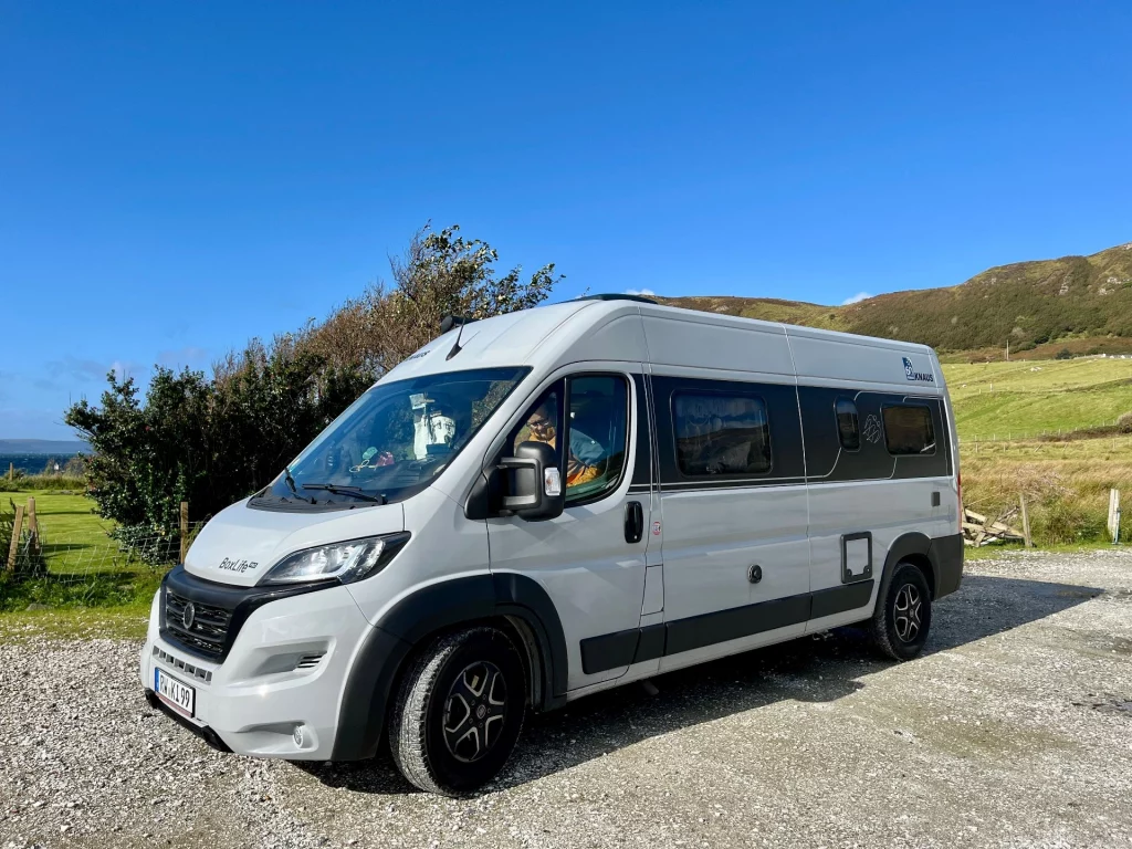 Seitenansicht eines hellgrauen Kastenwagens (Van) mit schwarzer Markise, der auf einem Schotterparkplatz vor einer grünen Hügellandschaft steht.