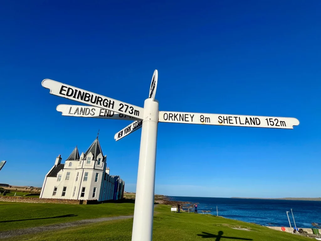Der weiße Wegweiser in John o’ Groats zeigt Entfernungen nach Edinburgh, Orkney und New York vor strahlend blauem Himmel.