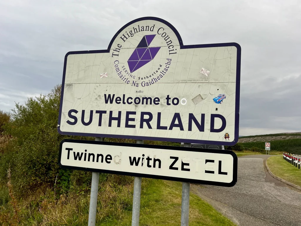 Ein wettergegerbtes weißes Ortsschild am Straßenrand in einer ländlichen, hügeligen Gegend Schottlands unter bewölktem Himmel. Das Hauptschild trägt das Logo des „Highland Council“ und die Aufschrift „Welcome to SUTHERLAND“ in großen, dunkelblauen Buchstaben. Darunter befindet sich ein kleineres Zusatzschild mit der Aufschrift „Twinned with ZETEL“. Das Schild weist Gebrauchsspuren wie Risse in der Oberfläche und einige kleine Aufkleber auf.