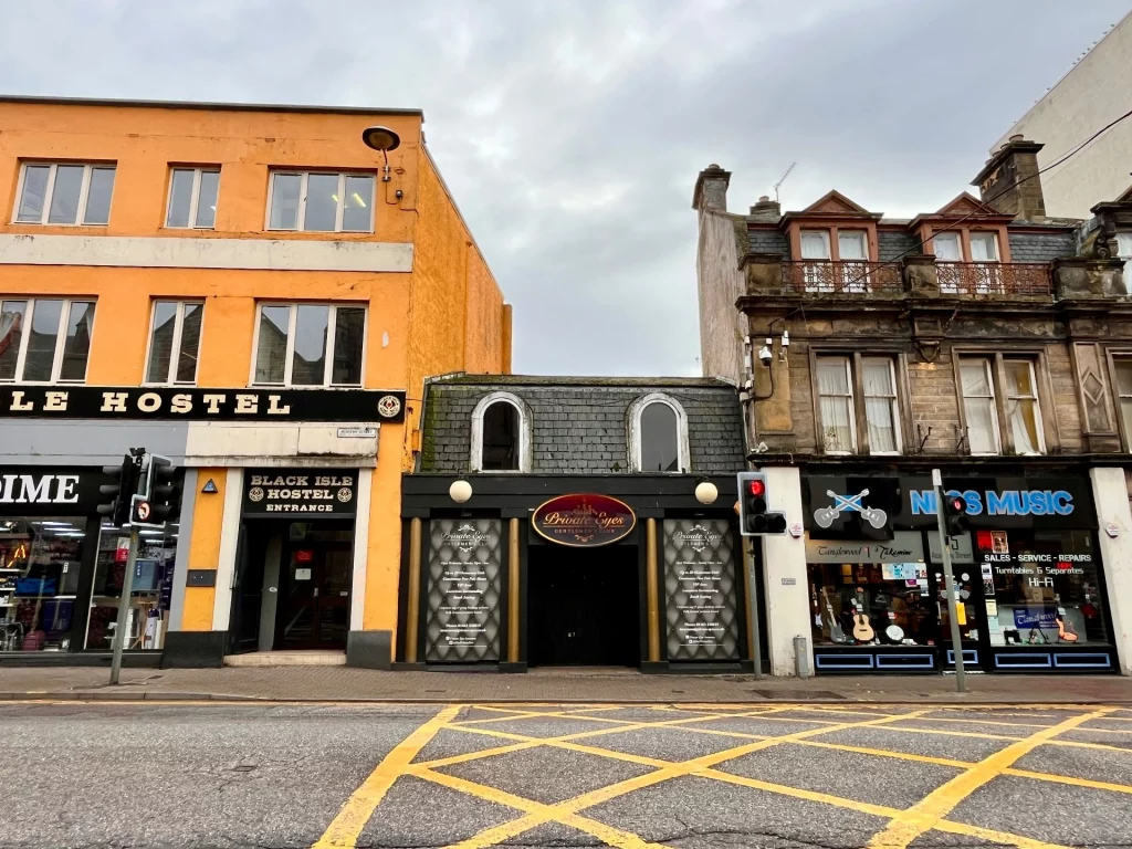 Eine Straßenansicht in Inverness mit drei unterschiedlichen Gebäuden: links ein markantes orangefarbenes Gebäude (Black Isle Hostel), mittig ein kleines dunkles Gebäude mit Mansarddach und rechts ein klassischer Altbau aus Sandstein.