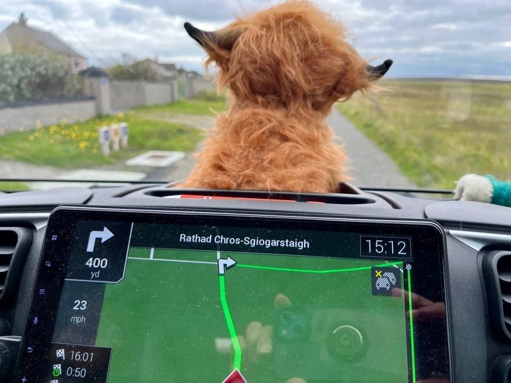 Blick über das Armaturenbrett eines Wohnmobils auf eine schmale Straße in Schottland. Im Vordergrund sitzt ein plüschiges Hochlandrind, im Display des Navigationsgeräts ist die Route nach Rathad Chros-Sgiogarstaigh zu sehen.