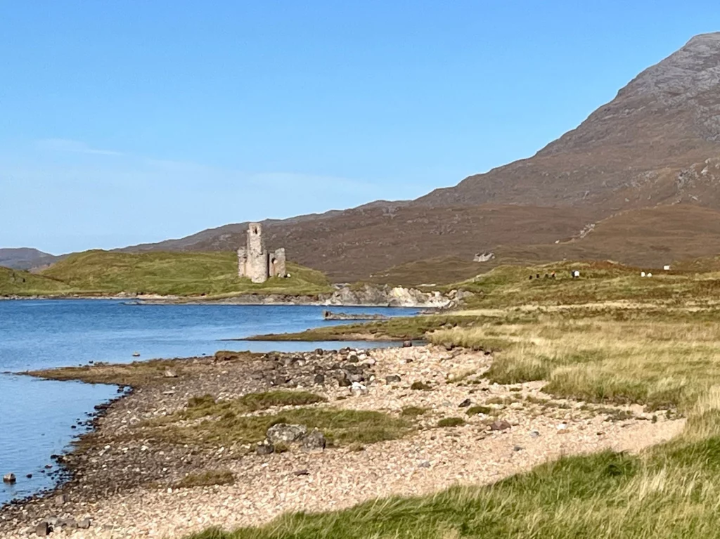 Die historische Ruine von Ardvreck Castle auf einer kleinen Halbinsel im Loch Assynt, umgeben von blauem Wasser und den Bergen der schottischen Highlands bei klarem Wetter.