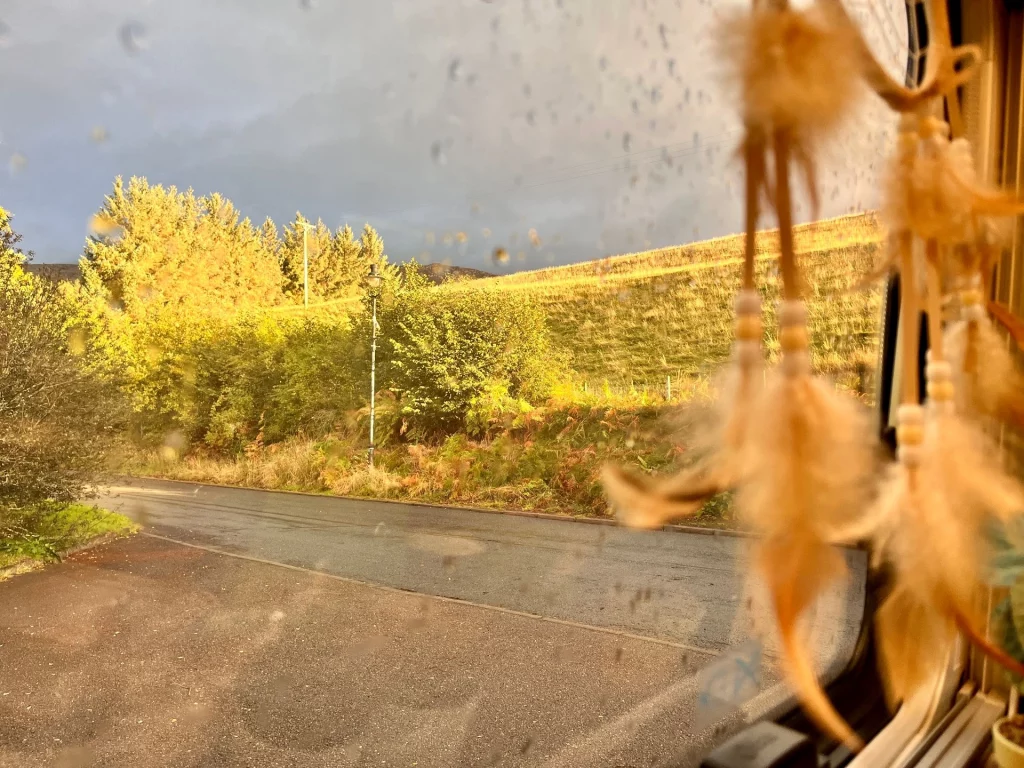 Blick durch ein regennasses Fenster eines Campers auf eine herbstliche schottische Landschaft. Im Vordergrund hängt ein Traumfänger mit Federn, im Hintergrund leuchten Bäume und ein Grashügel im warmen Sonnenlicht.