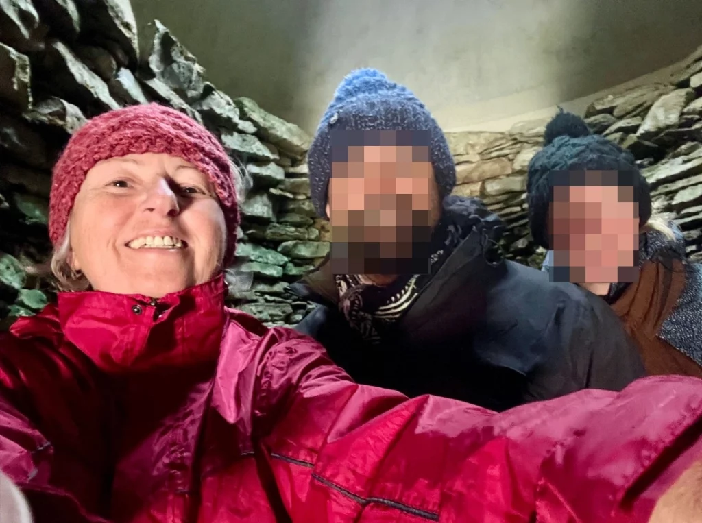 Ein Selfie von drei Personen in wetterfester Kleidung und Mützen im Inneren einer neolithischen Steinkammer. Im Hintergrund sind die charakteristischen, kunstvoll aufgeschichteten Trockenmauerwände des Cairns zu sehen.