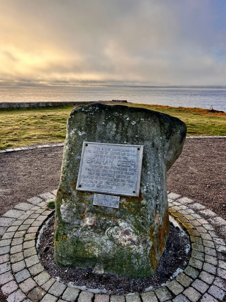 Ein massiver, mit Moos bewachsener Gedenkstein am Chanonry Point mit einer Metallplakette, die an den Brahan Seer erinnert. Im Hintergrund sind das Grasland und das ruhige Meer bei Sonnenaufgang zu sehen.