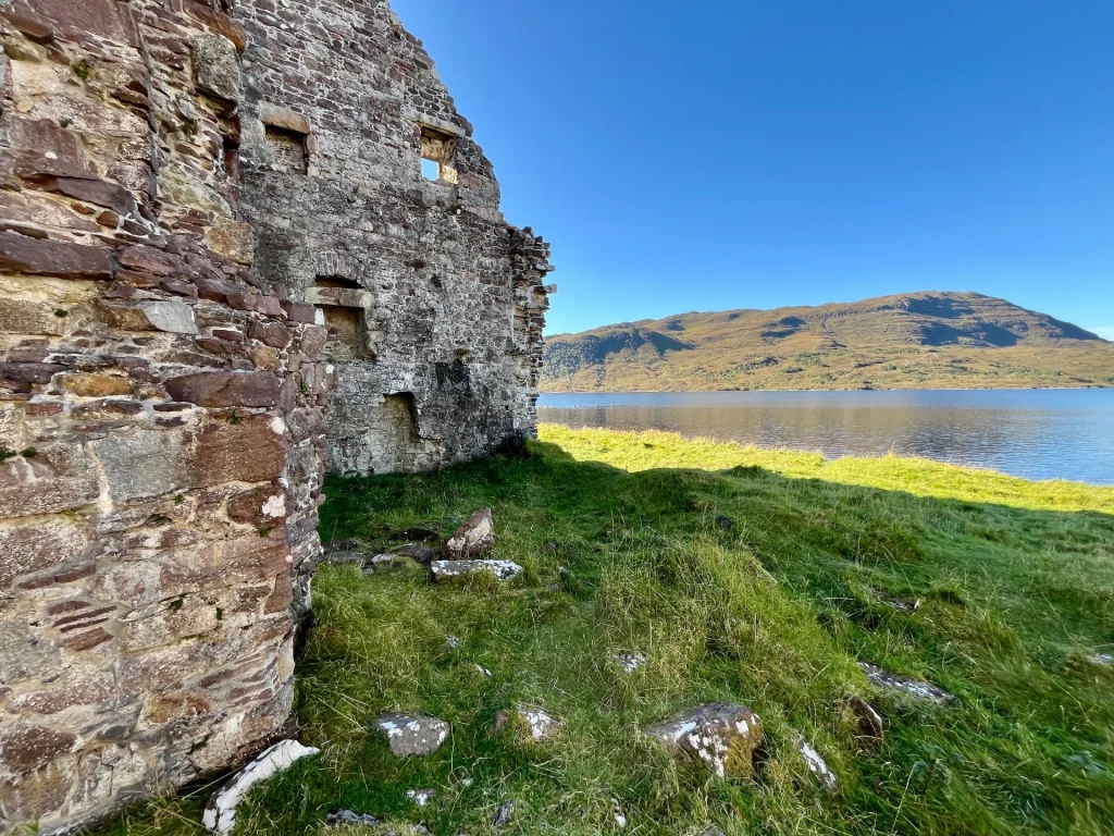 Nahaufnahme der steinernen Mauerreste der Calda House Ruine mit Blick auf den Loch Assynt und die gegenüberliegenden Berge unter blauem Himmel.