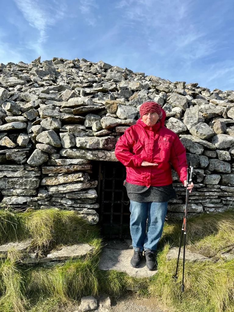 Eine Person in einer roten Jacke steht vor der massiven, aus grauen Steinen aufgeschichteten Wand eines neolithischen Grabhügels.