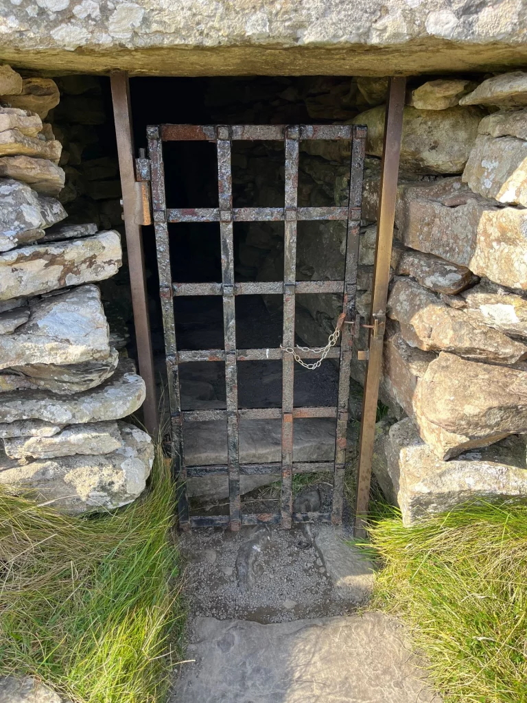Nahaufnahme eines niedrigen Eingangs in ein Steingrab, versperrt durch ein rostiges Metallgitter vor einem dunklen Gang.