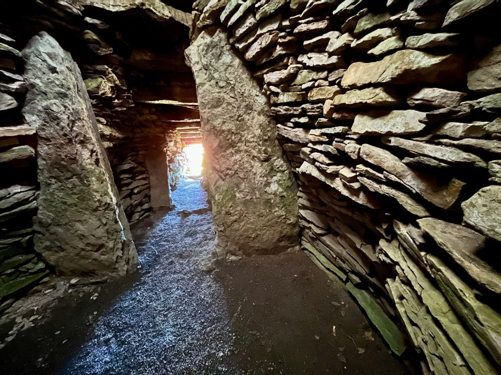 Innenansicht eines schmalen, niedrigen Ganges aus trocken gemauerten Steinwänden, an dessen Ende helles Tageslicht durch den Ausgang scheint.