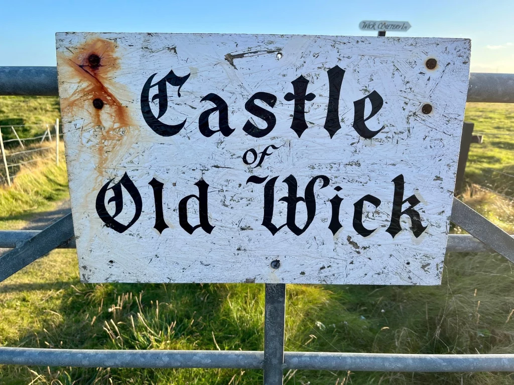 Ein weißes Holzschild mit der Aufschrift "Castle of Old Wick" in schwarzer Frakturschrift vor einer Wiese.
