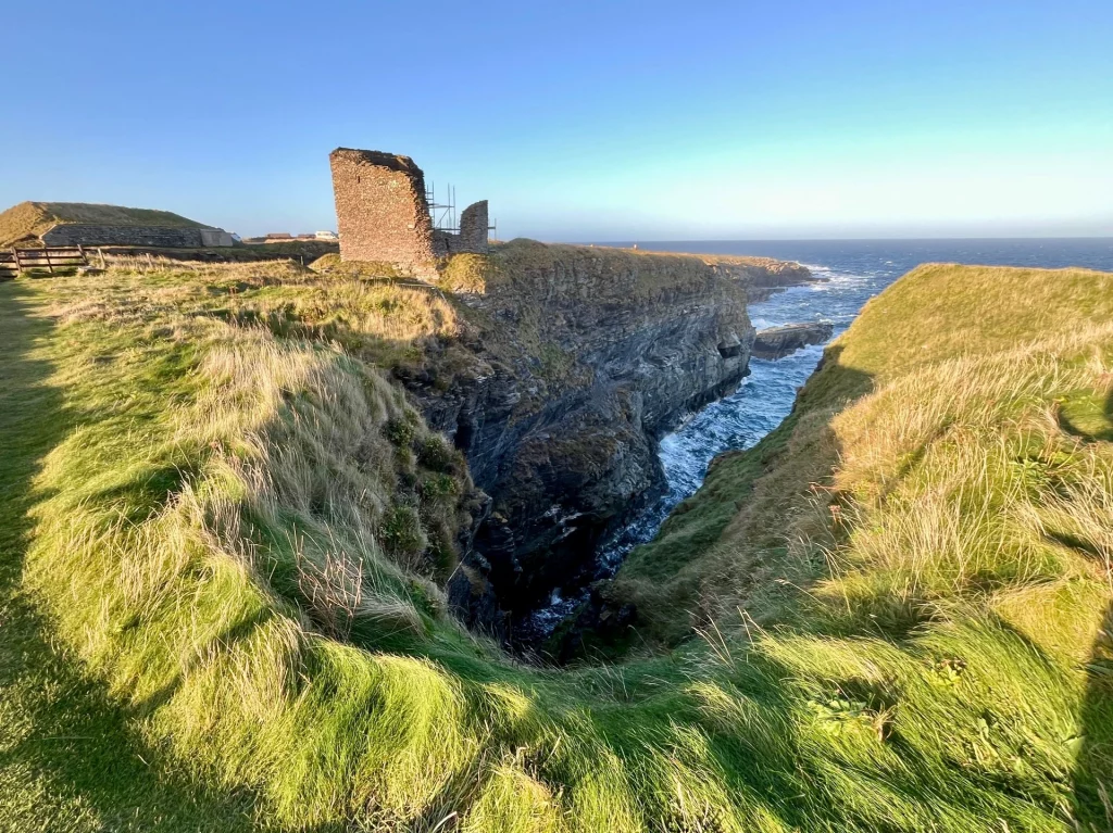 Weitwinkelaufnahme einer tiefen Bucht im Fels mit dem einsamen Turm des Castle of Old Wick im Hintergrund.