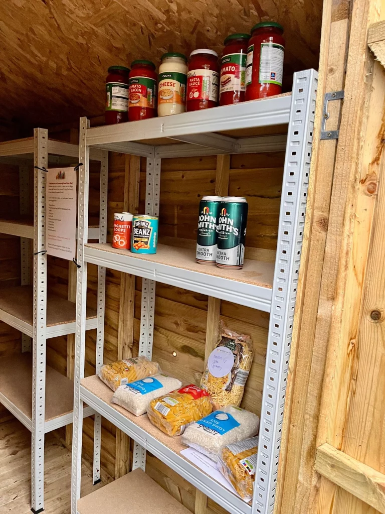Das Innere einer Holzhütte mit Metallregalen. In den Regalen stehen verschiedene Lebensmittel wie Tomatensauce, Bohnen, Nudeln, Reis und zwei Dosen John Smith’s Bier.