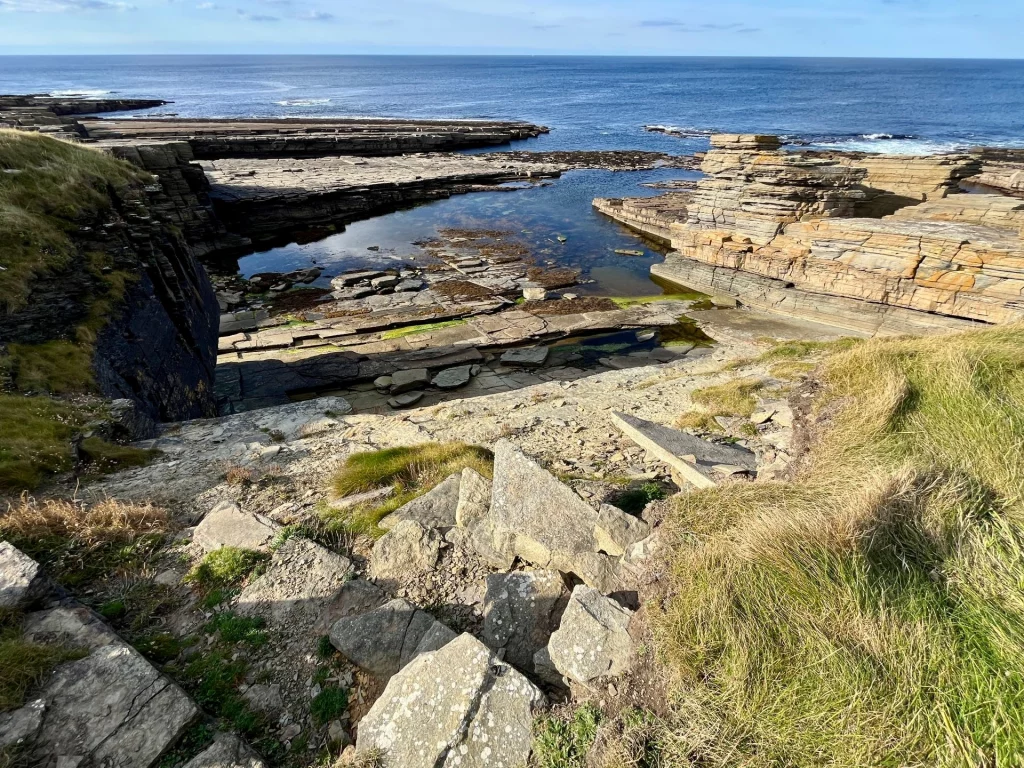 Eine dramatische Küstenansicht von oben herab auf geschichtete Felsplateaus, die ins tiefblaue Meer ragen. Im Vordergrund sind steile, grasbewachsene Hänge und lose Felsbrocken zu sehen, während sich im Wasser flache, dunkle Gesteinsformationen abzeichnen.