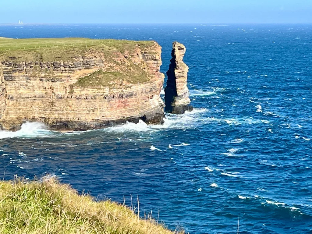 Eine markante, alleinstehende Felsnadel direkt an der Abbruchkante einer hohen Klippe, im Hintergrund die Pentland Skerries.