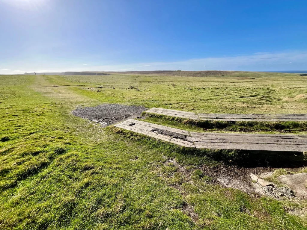 Ein grüner Wanderweg auf einem Hochplateau an der schottischen Küste unter strahlend blauem Himmel mit Holzstegen im Vordergrund.