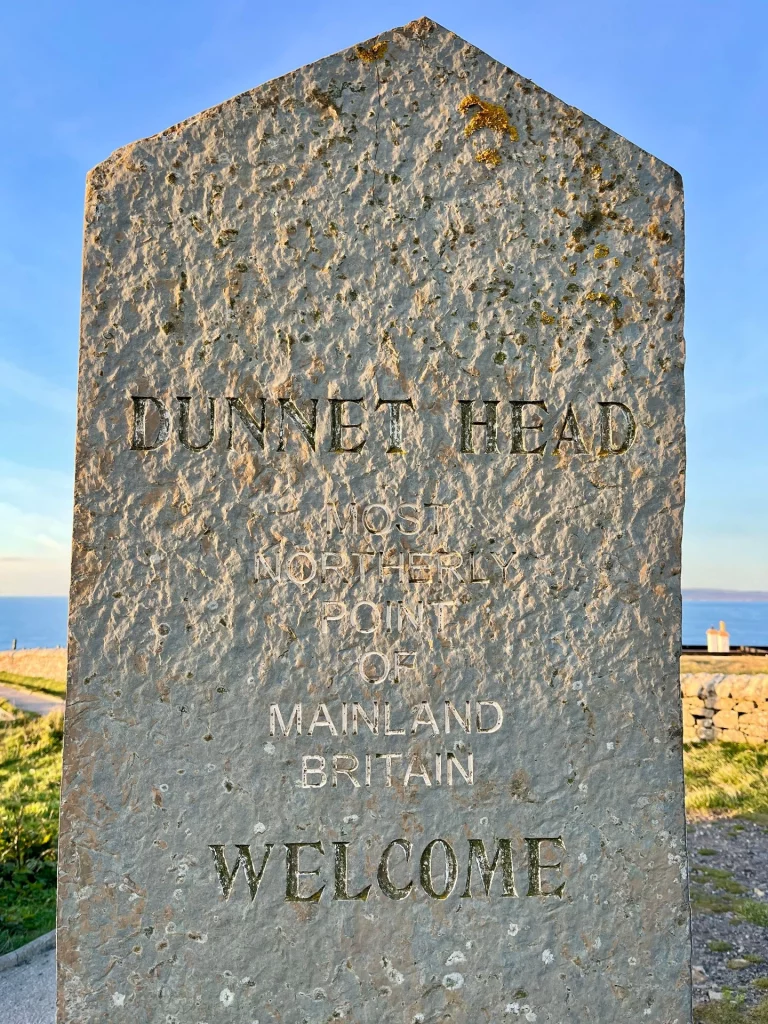 Nahaufnahme eines grauen Steingedenksteins mit der eingravierten Aufschrift: „DUNNET HEAD - MOST NORTHLY POINT OF MAINLAND BRITAIN - WELCOME“. Der Stein ist leicht verwittert und steht vor einem Hintergrund aus blauem Himmel.