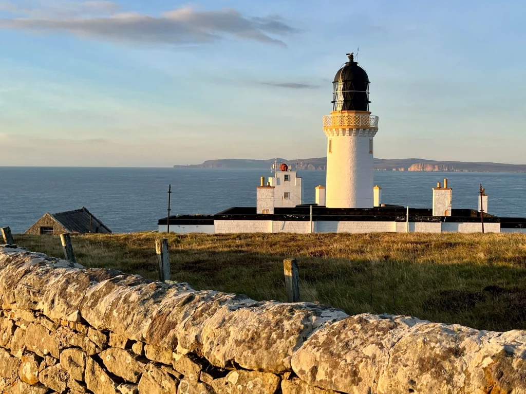 Panoramaaufnahme des Dunnet Head Leuchtturms bei warmem Abendlicht. Der weiße Turm mit schwarzer Kuppel steht hinter einer alten Trockenmauer auf einer Klippe. Im Hintergrund erstreckt sich das ruhige Meer bis zu den fernen Klippen der Orkney-Inseln am Horizont.