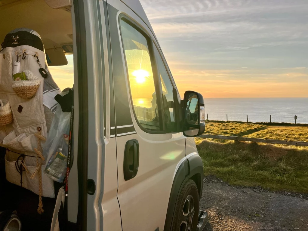 Blick aus der offenen Schiebetür eines weißen Kastenwagens. Im Fenster spiegelt sich die tiefstehende, goldene Sonne. Draußen erstreckt sich eine grüne Klippenlandschaft, die im sanften Abendlicht bis zum ruhig daliegenden Meer führt. Im Inneren des Vans ist eine praktische Sitztaschen-Aufbewahrung an der Kopfstütze erkennbar.