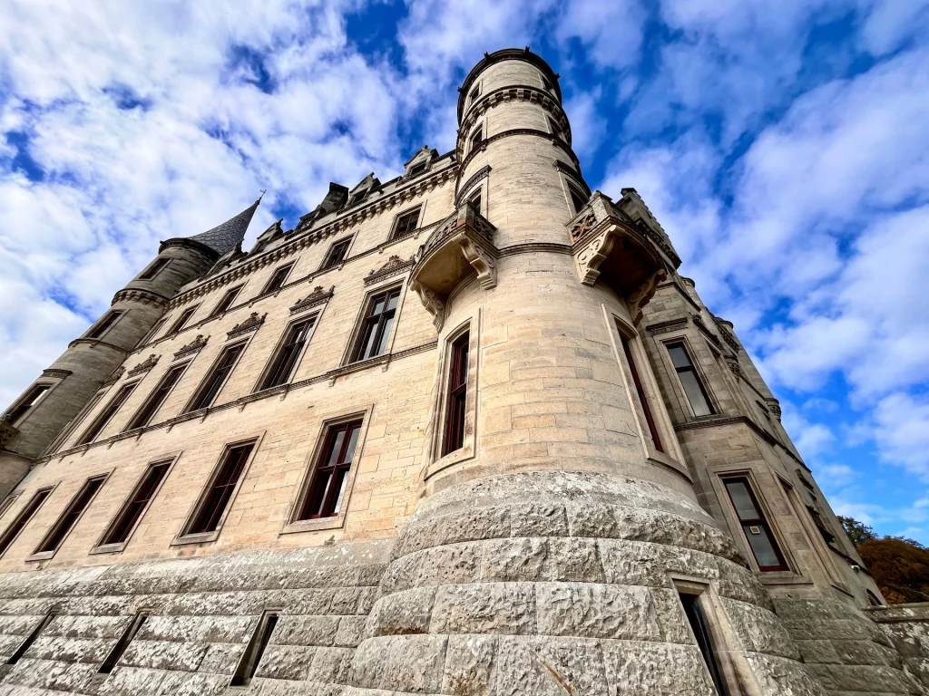 Eine dramatische Untersicht der hellen Steinfassade von Dunrobin Castle. Zu sehen sind ein runder Ecktorturm mit kleinen Balkonen, zahlreiche Fensterreihen und ein spitzer Turmhelm gegen einen blau-weißen Wolkenhimmel.