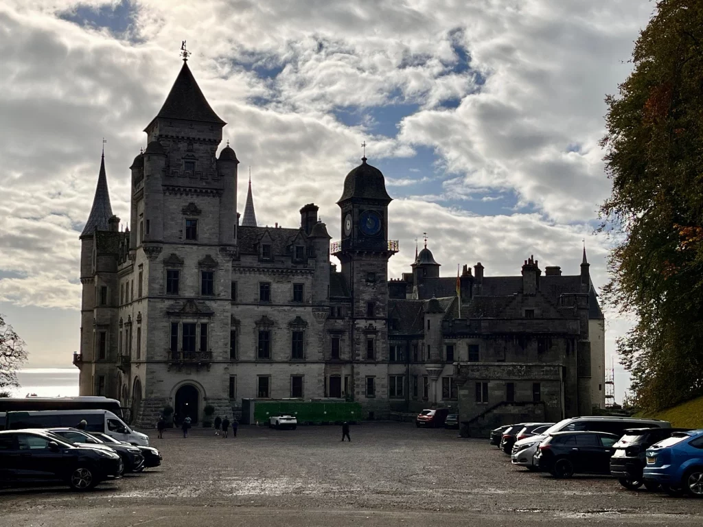 Eine Aufnahme von Dunrobin Castle von der Hofseite aus. Das graue Steinschloss zeigt verschiedene Türme, darunter einen markanten Uhrturm und spitze Turmhelme. Im Vordergrund befindet sich ein geschotterter Parkplatz mit mehreren geparkten Autos und Besuchern vor der dramatischen Kulisse eines bewölkten Himmels.