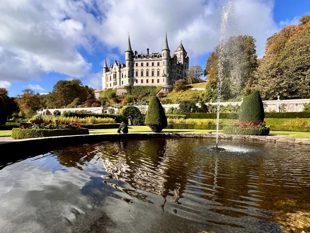 Das majestätische Dunrobin Castle auf einer Anhöhe, davor ein kreisrunder Springbrunnen im Vordergrund, der sich im Wasser spiegelt. Gepflegte Gartenanlagen und herbstliche Bewaldung umgeben das Schloss.