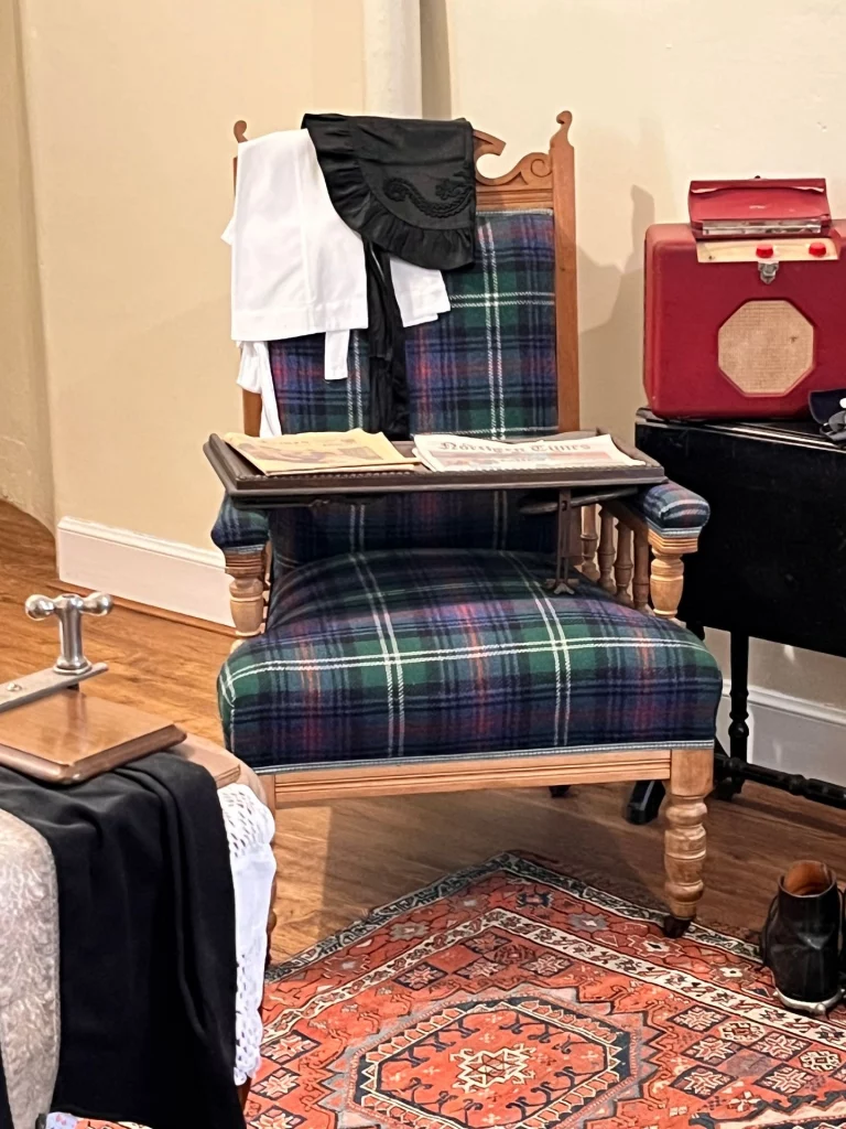 Ein klassischer Holzstuhl mit Armlehnen, dessen Polsterung komplett in einem grün-blau-rot karierten Tartan-Muster gehalten ist. Auf dem Stuhl liegt ein hölzernes Tablett mit Zeitungen, im Hintergrund sind historische Gegenstände wie ein roter Koffer und Kleidung zu sehen.