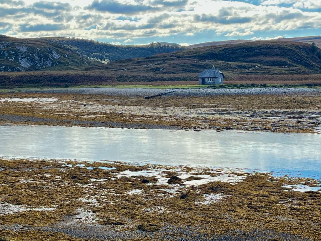 Ein einsames, graues Steinhäuschen (Bothy) an einem Meeresarm bei Kylesku in Schottland vor einer Kulisse aus dramatischen Wolken und kargen Hügeln.