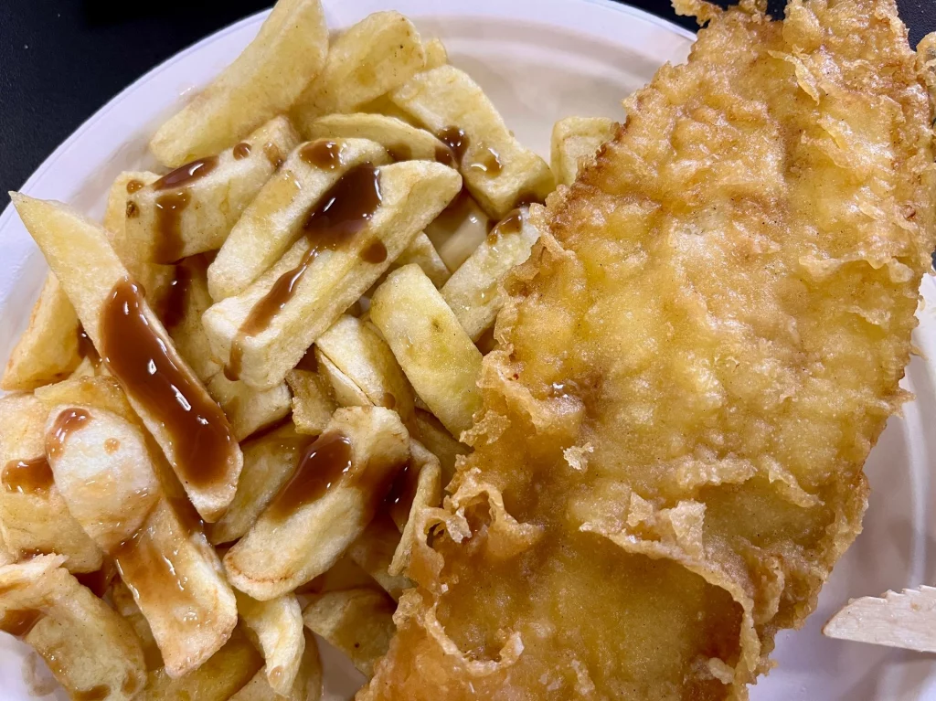 Eine Portion goldbraun gebackener Fisch mit Pommes (Chips) und brauner Sauce auf einem Teller.