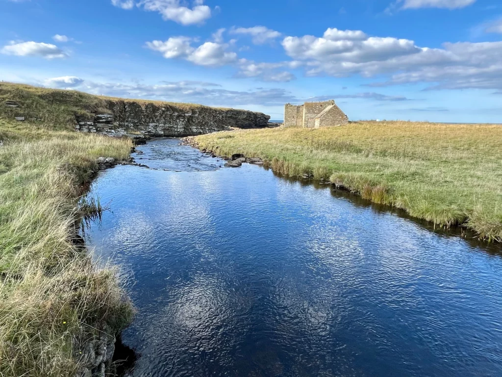 Ein sanft geschwungener, blauer Fluss fließt durch eine flache Graslandschaft unter einem sonnigen Himmel. Im Hintergrund ist ein einzelnes, steinernes Gebäude am Ufer zu sehen.
