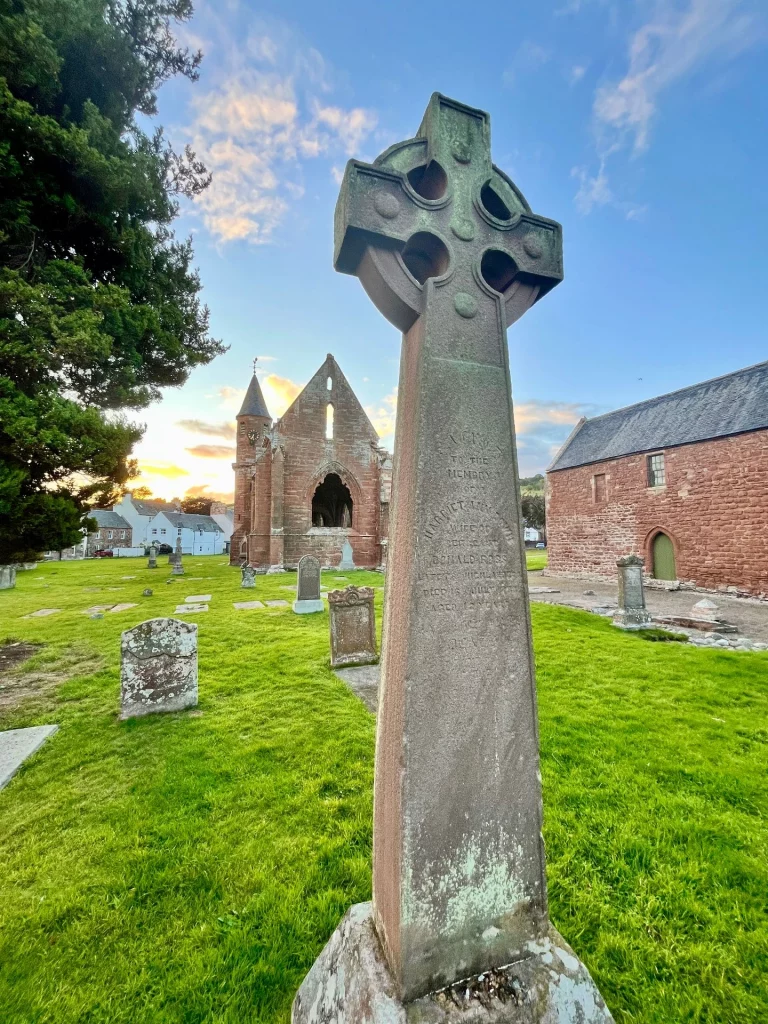 Ein hohes keltisches Steinkreuz im Vordergrund eines grünen Friedhofs bei Sonnenuntergang. Im Hintergrund ist die Ruine der Fortrose Cathedral aus rotem Sandstein unter einem blau-gelben Himmel zu sehen.