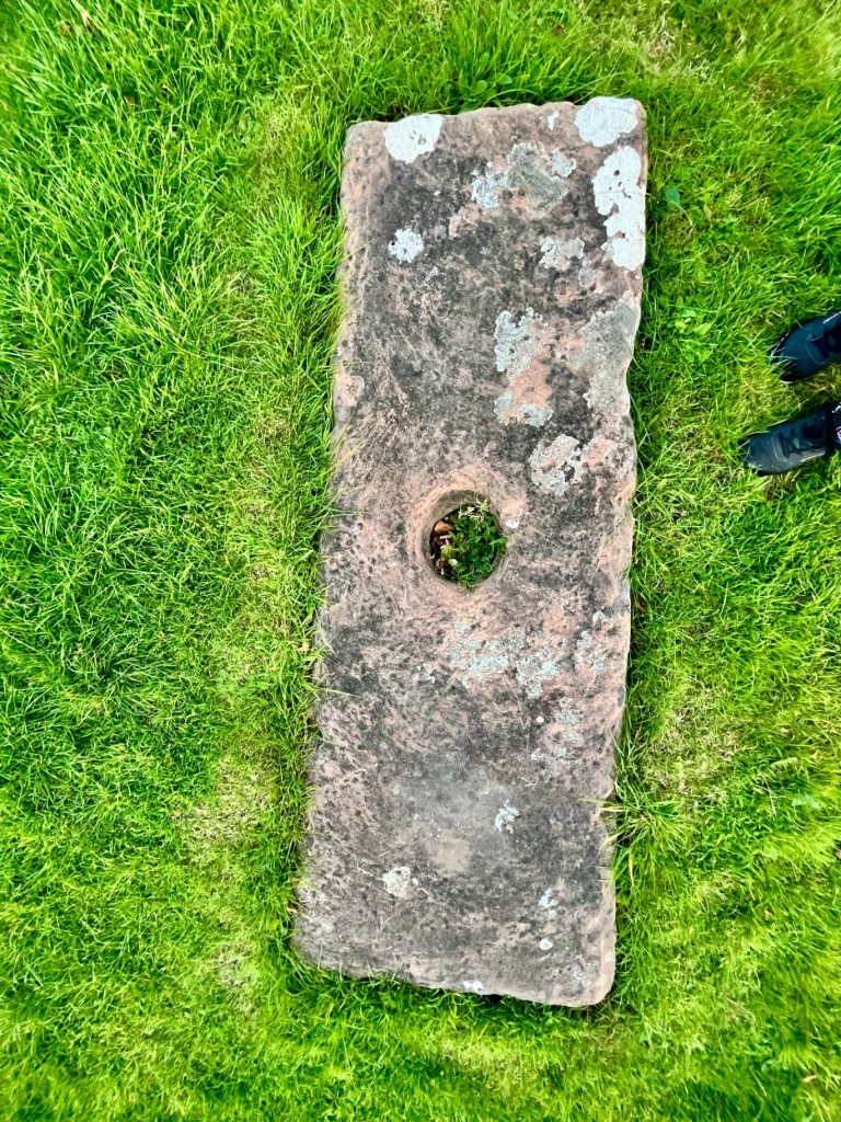 Draufsicht auf eine flache, rechteckige Steinplatte im Gras mit einem kreisrunden Loch in der Mitte. Moos und Flechten bedecken die verwitterte Oberfläche des rötlichen Gesteins.
