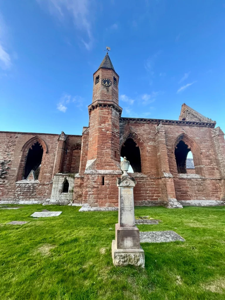 Die beeindruckende Ruine der Fortrose Cathedral mit ihrem markanten achteckigen Glockenturm und gotischen Fensterbögen. Ein weißes Denkmal steht auf dem gepflegten Rasen im Vordergrund.