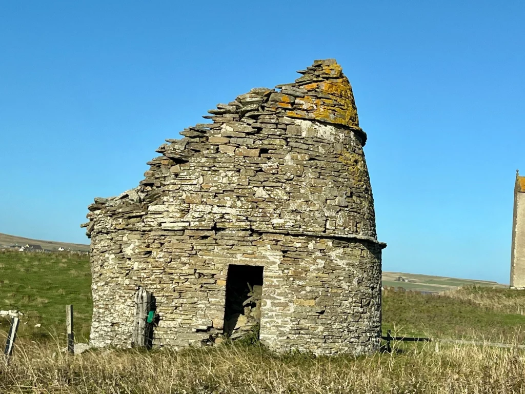 Eine Bienenkorb-förmige Ruine eines historischen Taubenturms aus geschichtetem Stein auf einer grünen Wiese unter blauem Himmel.